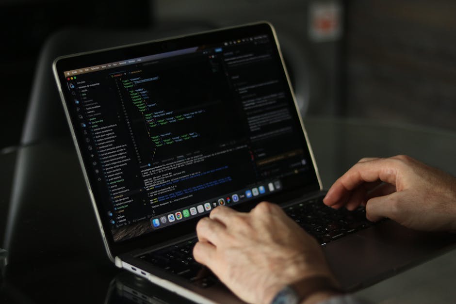 A close-up view of a person's hands working on a laptop placed on a dark workspace, with the screen displaying lines of code and a dark theme interface. The individual's fingers are positioned over th